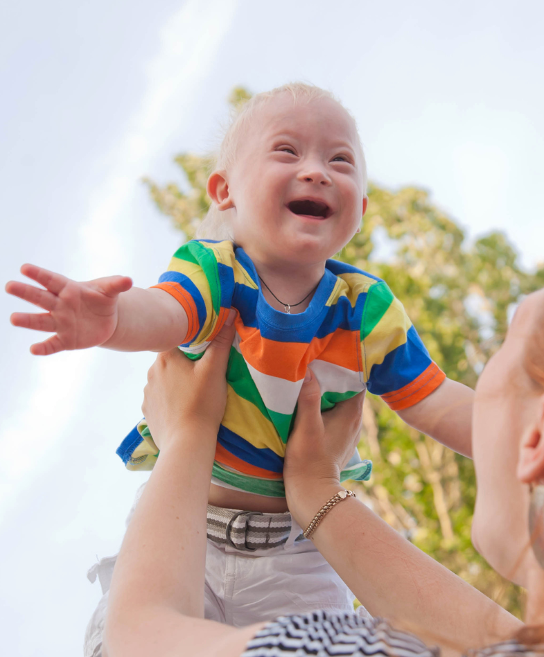 A young child with Down Syndrome smiling while being held up in the air by an adult